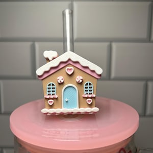 Puede incluir: Un adorno para pajita con forma de casa de jengibre en tonos marrón, rosa y blanco, con una puerta azul y una pajita transparente. El adorno está colocado sobre una tapa rosa, lo que sugiere un recipiente para bebidas. El diseño incluye corazones y caramelos.