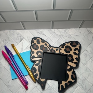 Puede incluir: Un marco decorativo en forma de lazo con un centro negro y un diseño de estampado de leopardo. Tres bolígrafos en rosa, morado y dorado están junto a un cuadrado de papel azul. El marco está sobre una superficie con vetas de mármol.