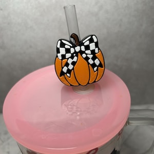 Op de afbeelding: Een doorzichtige rietjes-topper in de vorm van een oranje pompoen met een zwart-wit geblokte strik. De topper is bevestigd aan een transparant rietje dat door een doorschijnend roze deksel op een glazen beker met een decoratief patroon steekt.