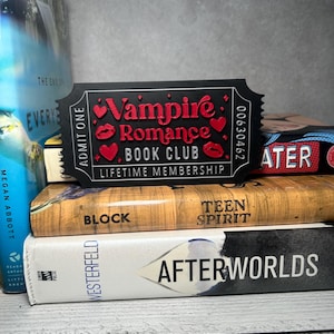 May include: A black ticket-shaped item with the words "Vampire Romance Book Club" in red, featuring red heart and lip graphics. The ticket reads "Lifetime Membership" and is placed on a stack of books.