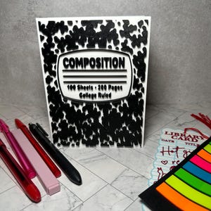 Puede incluir: Un cuaderno blanco con un patrón de manchas negras y una etiqueta central que dice COMPOSITION 100 Sheets 200 Pages College Ruled. Junto a él hay cuatro bolígrafos en rojo, rosa y negro, y un marcapáginas de biblioteca con pestañas de colores.