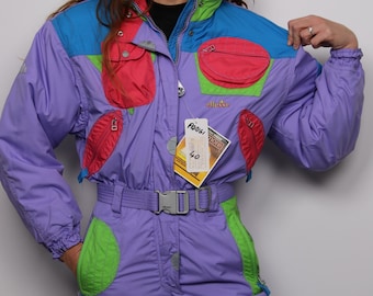 May include: A purple ski suit with blue, green, and pink accents. The suit has a zipper closure, a belt, and multiple pockets. The suit has a tag that says "Preis! 40".