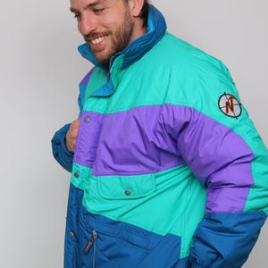 Puede incluir: Una chaqueta acolchada multicolor con un esquema de color azul, verde y morado. La chaqueta tiene una cremallera y un bolsillo grande en la parte delantera. La chaqueta tiene un logotipo en la manga derecha que dice "North Face" con un diseño de brújula.