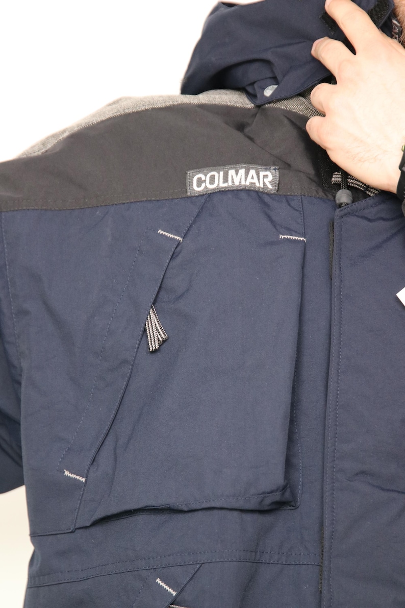 May include: A navy blue jacket with a black and gray panel across the shoulders. The jacket has a zipper closure and a pocket on the left side. The word "COLMAR" is printed in white on a black patch on the jacket.