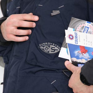 May include: A navy blue ski jacket with a black collar and a zipper closure. The jacket has a large pocket on the left side with the text "OVERSYSTEM" and a logo. The jacket also has a smaller pocket on the right side with a zipper closure. The jacket is made of a water-resistant material and is designed for cold weather.