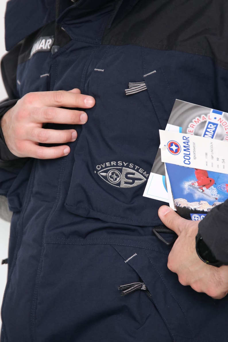 May include: A navy blue ski jacket with a black collar and a zipper closure. The jacket has a large pocket on the left side with the text "OVERSYSTEM" and a logo. The jacket also has a smaller pocket on the right side with a zipper closure. The jacket is made of a water-resistant material and is designed for cold weather.