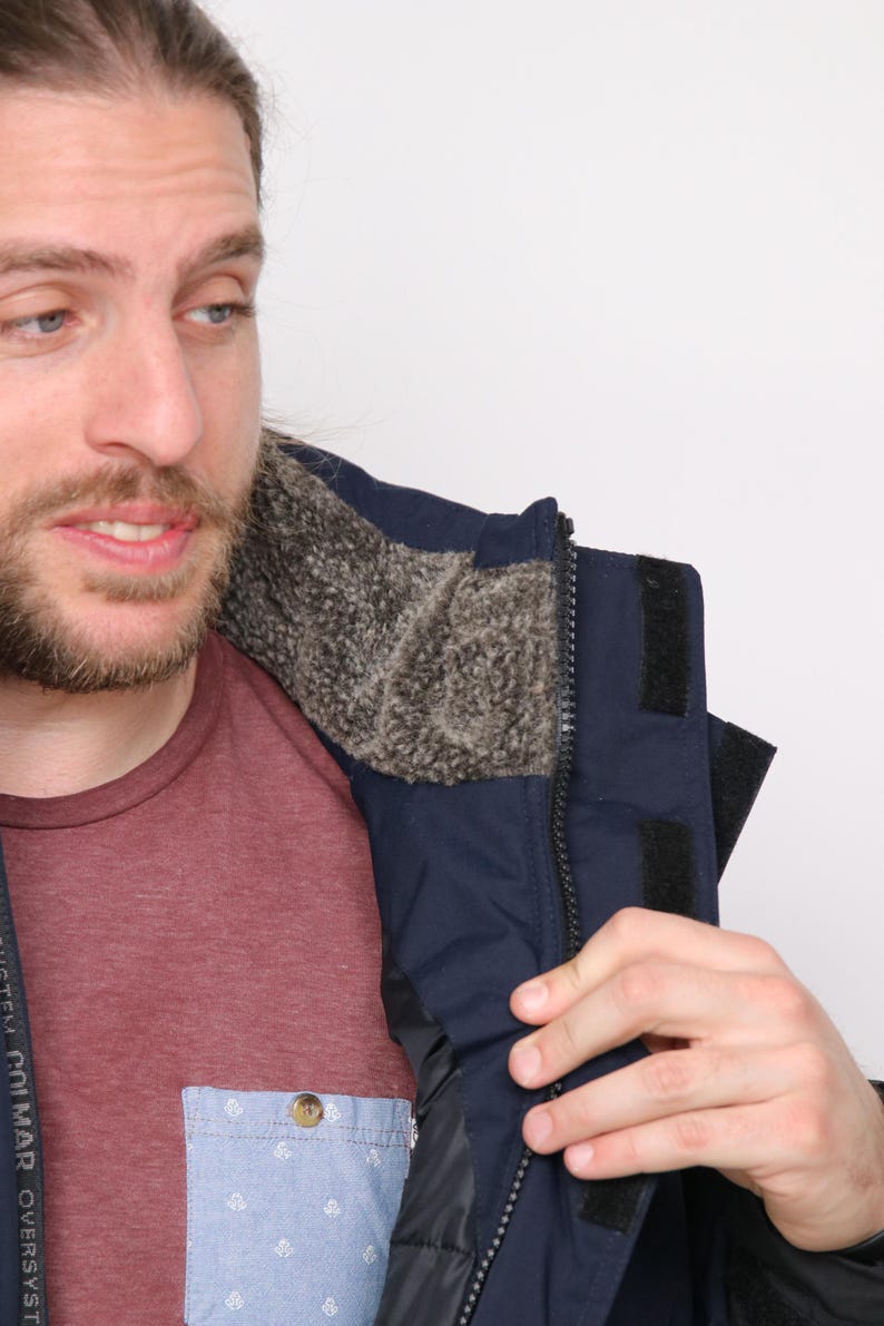 May include: A navy blue jacket with a black zipper and a fleece lining. The jacket has a velcro strap on the left side of the collar. The text "SYSTEM CO. MAR. OVERSYST" is visible on the left side of the jacket.