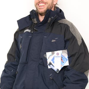 May include: A navy blue and gray ski jacket with a black zipper and a Colmar logo. The jacket has a hood and multiple pockets. The jacket has a tag with the Colmar logo and the text "Colmar" on it.