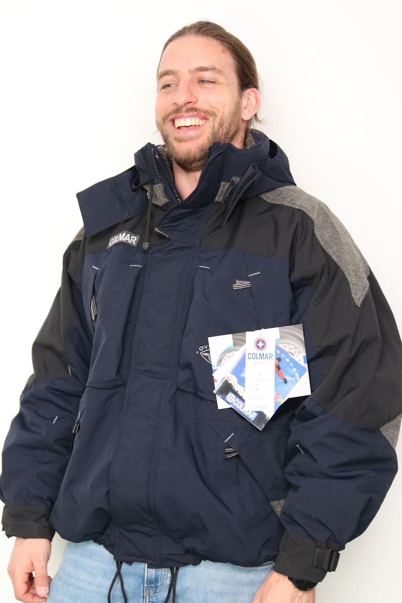 May include: A navy blue and gray ski jacket with a black zipper and a Colmar logo. The jacket has a hood and multiple pockets. The jacket has a tag with the Colmar logo and the text "Colmar" on it.