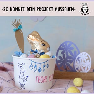 Puede incluir: Un vaso de papel blanco con un dise&ntilde;o de conejo y el texto "Frohe Ostern" (Feliz Pascua) est&aacute; lleno de huevos de chocolate, un conejito de chocolate y una zanahoria de papel marr&oacute;n con una cinta azul. El vaso est&aacute; sobre un mantel a cuadros rosa y blanco.