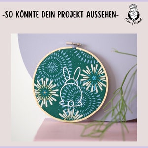 K&ouml;nnte beinhalten: Ein gr&uuml;ner Stickrahmen mit einer wei&szlig;en Umrandung eines Kaninchens auf einem gr&uuml;n-blauen Blumenmuster.