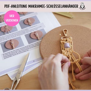 Może przedstawiać: Zbliżenie na breloczek z makramą z jasnofioletową koralikiem. Breloczek jest trzymany przez rękę i leży na powierzchni korkowej. Obraz zawiera instrukcje tworzenia breloczka, z widocznym tekstem "PDF-ANLEITUNG MAKRAMEE-SCHLÜSSELANHÄNGER" i "VIER VERSIONEN".