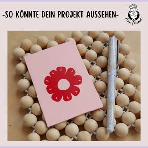 Könnte beinhalten: Ein rosa Notizbuch mit rotem Blumenmuster und ein Stift auf einer Holzperlenmatte. Der Text "-SO KÖNNTE DEIN PROJEKT AUSSEHEN-" steht oben.
