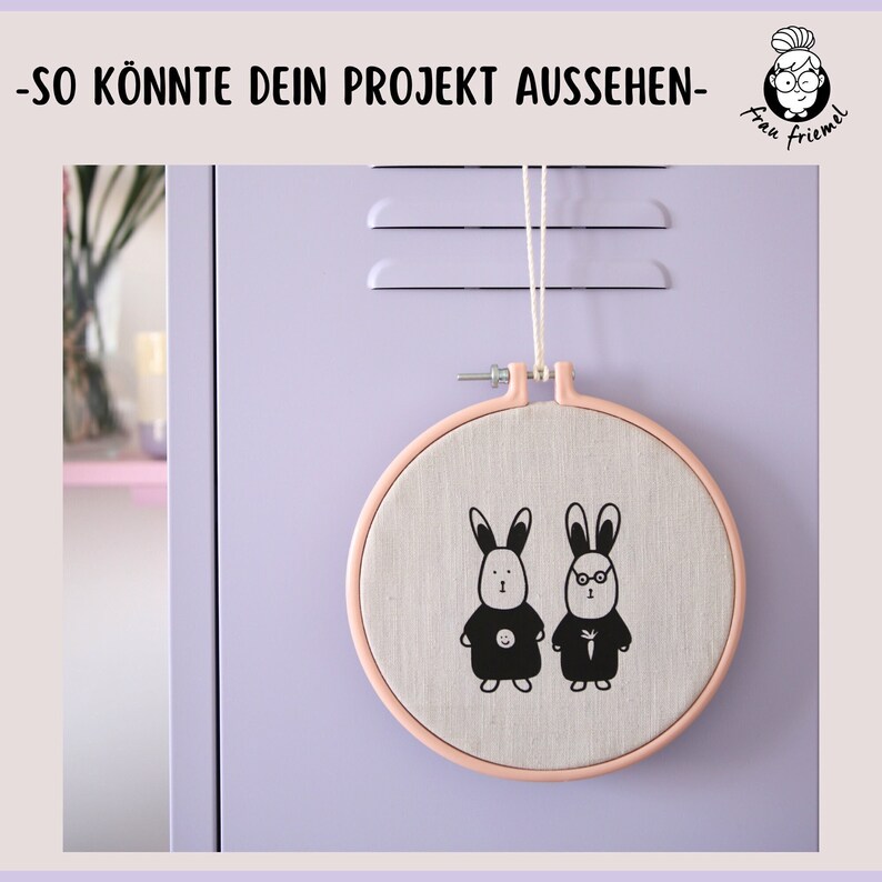 K&ouml;nnte beinhalten: Ein rosa Stickrahmen mit einem beigefarbenen Stoff im Inneren. Der Stoff zeigt eine schwarz-wei&szlig;e Illustration von zwei Hasen mit einer Karotte. Der Text "So k&ouml;nnte dein Projekt aussehen" befindet sich oben im Bild.