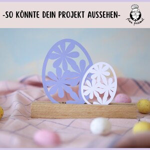 Puede incluir: Dos huevos de Pascua de papel con dise&ntilde;os de flores recortados. El huevo m&aacute;s grande es morado y el m&aacute;s peque&ntilde;o es blanco. Los huevos se muestran en un soporte de madera.