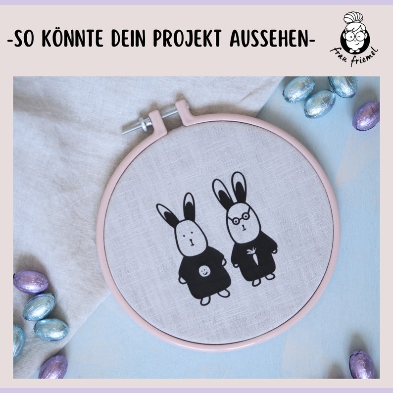 K&ouml;nnte beinhalten: Ein rosa Stickrahmen mit wei&szlig;em Leinenstoff im Inneren. Der Stoff zeigt zwei schwarz-wei&szlig;e Hasenillustrationen. Ein Hase tr&auml;gt eine Brille und der andere hat ein Smiley auf seinem Shirt. Der Text "Frau Friemel" befindet sich in der oberen rechten Ecke des Bildes.