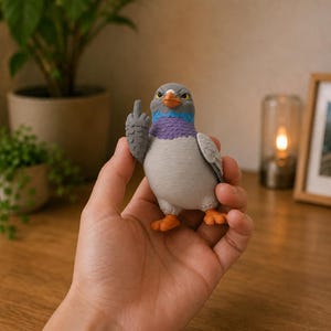 Hand holding a humorous pigeon figurine to demonstrate its scale, with layered feather details, bright neck colors and a cheeky hand gesture.