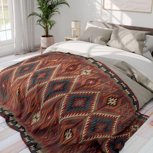 May include: A colorful patterned blanket with a geometric design in shades of red, orange, blue, and beige covers a bed. The blanket has a textured appearance and is draped over the bed, showcasing its intricate pattern. The bed is made with white and gray bedding.