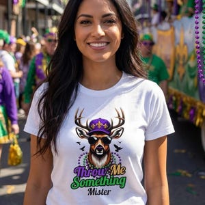 May include: White t-shirt featuring a deer graphic wearing sunglasses and a purple cap. The text "Throw Me Something Mister" is below the deer. The image is taken outdoors with people in the background.