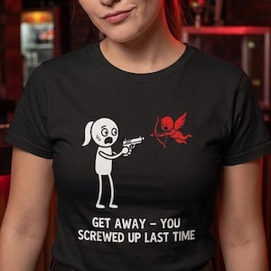 Puede incluir: Camiseta negra con un gráfico humorístico de un muñeco de palo apuntando con una pistola a un cupido rojo. El texto "GET AWAY - YOU SCREWED UP LAST TIME" está impreso debajo.