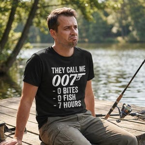 May include: Black t-shirt with the text "THEY CALL ME 007 - 0 BITES - 0 FISH - 7 HOURS" in white. The shirt is worn by a person sitting on a wooden dock with a fishing rod.