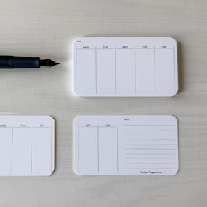 May include: A dark blue fountain pen rests near three white weekly planner notepads. Each notepad has a rounded rectangle shape with days of the week printed on them. One notepad has lined notes section.