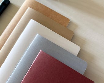 May include: A stack of five rectangular notebooks with paper covers in shades of brown, cream, light gray, dark gray, and deep red, arranged in a diagonal overlapping pattern next to a black pen and a dark ink bottle.