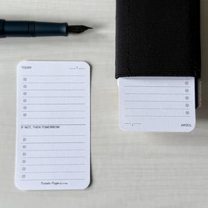 May include: A black fountain pen rests near two white notepads with rounded corners. One notepad is loose, the other is in a black elastic holder. The notepads have lines and checkboxes, with the text "TODAY" and "IF NOT, THEN TOMORROW".