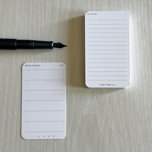 May include: Two white pocket-sized notepads with rounded corners, one labeled "Weekly Repeats" and the other "Notes/Review." A black fountain pen is positioned above the notepads. The notepads have lined and grid paper.