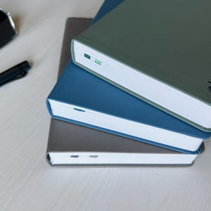 Three PU-leather notebooks: gray, blue, and green are stacked on top of each other on a white wooden desk. There is a fountain pen and an ink bottle placed next to it.