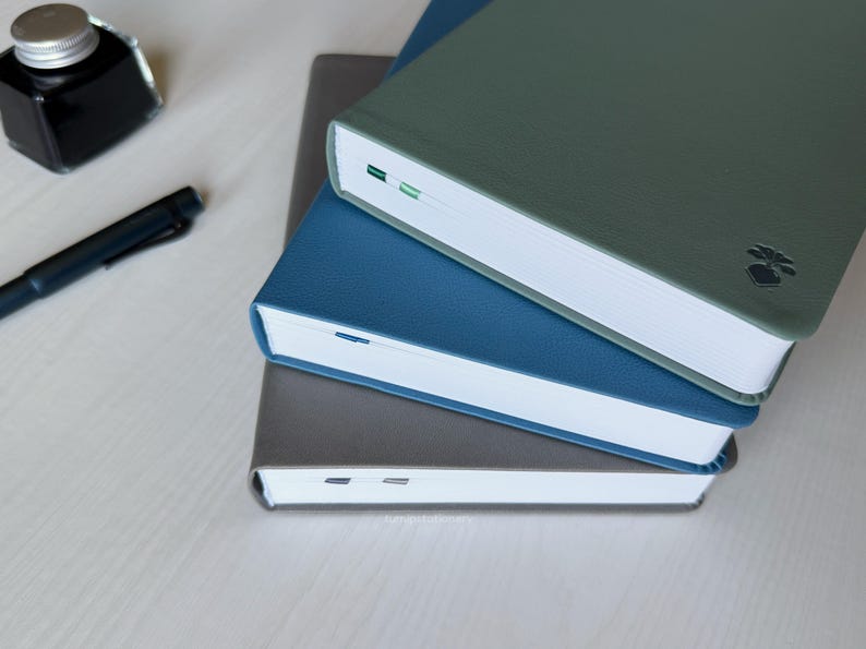 Three PU-leather notebooks: gray, blue, and green are stacked on top of each other on a white wooden desk. There is a fountain pen and an ink bottle placed next to it.