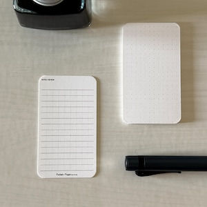 A set of business card-sized note cards on a white table. The card on the left has a line with mini vertical dots layout with the text "Pockets + Pages by Turnip" at the bottom. The stack of cards on the right has a dot grid layout. A black ink bottle and small blue pen is placed nearby.