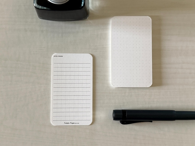 A set of business card-sized note cards on a white table. The card on the left has a line with mini vertical dots layout with the text "Pockets + Pages by Turnip" at the bottom. The stack of cards on the right has a dot grid layout. A black ink bottle and small blue pen is placed nearby.