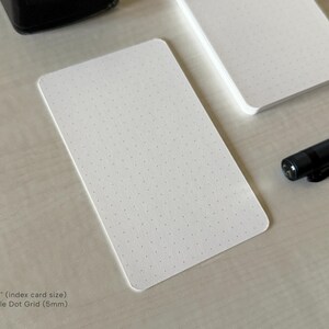 A side view of a set of index card-sized, dot grid note cards on a white table. A black ink bottle and small blue pen is placed nearby. Additional text on the bottom left in the image provides more details on the product including the size and layout type.