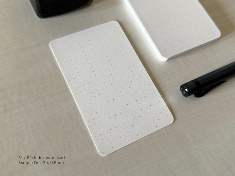 A side view of a set of index card-sized, dot grid note cards on a white table. A black ink bottle and small blue pen is placed nearby. Additional text on the bottom left in the image provides more details on the product including the size and layout type.