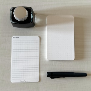 A set of index card-sized, note cards on a white table. The card on the left has a line with mini vertical dots layout with the text "Pockets + Pages by Turnip" at the bottom. The stack of cards on the right has a dot grid layout. A black ink bottle and small blue pen is placed nearby.