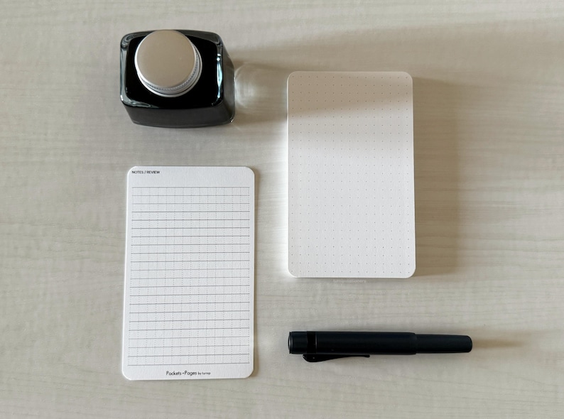 A set of index card-sized, note cards on a white table. The card on the left has a line with mini vertical dots layout with the text "Pockets + Pages by Turnip" at the bottom. The stack of cards on the right has a dot grid layout. A black ink bottle and small blue pen is placed nearby.