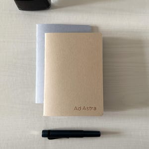 May include: Two notebooks, one silver and one tan, with the text "Ad Astra" on the tan notebook. A black pen and a black object are also present on a light-colored wooden surface.