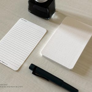 A side view of a set of index card-sized, note cards on a white table. The card on the left has a line with mini vertical dots layout with the text "Pockets + Pages by Turnip" at the bottom. The stack of cards on the right has a dot grid layout. A black ink bottle and small blue pen is placed nearby.