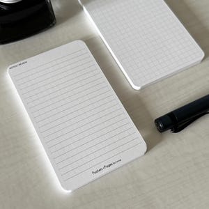 A side view of a set of index card-sized note cards on a white table. The card on the left has a line with mini vertical dots layout with the text "Pockets + Pages by Turnip" at the bottom. The stack of cards on the right has a lightly printed graph layout. A black ink bottle and small blue pen is placed nearby.