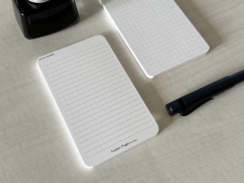 A side view of a set of index card-sized note cards on a white table. The card on the left has a line with mini vertical dots layout with the text "Pockets + Pages by Turnip" at the bottom. The stack of cards on the right has a lightly printed graph layout. A black ink bottle and small blue pen is placed nearby.