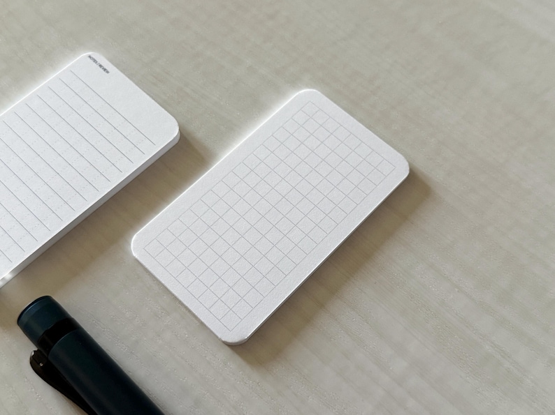 A side view of a set of business card-sized graph cards on a white table, next to a small stack of line note cards. A small blue pen is placed nearby.