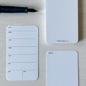 May include: A dark blue fountain pen rests above three white notepads. One notepad is a weekly planner, another is blank, and the third has the text "Notes". The planner has lines for each day of the week.