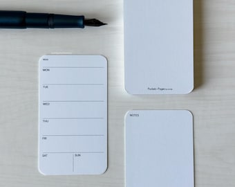 May include: A dark blue fountain pen rests above three white notepads. One notepad is a weekly planner, another is blank, and the third has the text "Notes". The planner has lines for each day of the week.