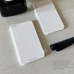 A side view of a set of business card-sized, note cards on a white table. The card on the left has a line with mini vertical dots layout with the text "Pockets + Pages by Turnip" at the bottom. The stack of cards on the right has a light graph layout. A black ink bottle and small blue pen is placed nearby. Additional text on the upper left in the image provides more details on the product including the size, material, and layout description.