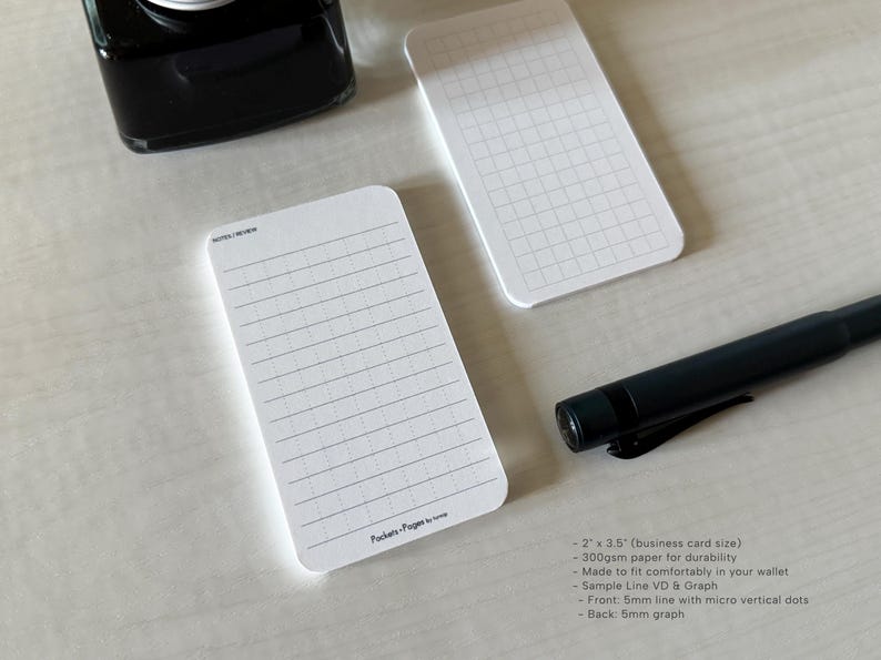 A side view of a set of business card-sized, note cards on a white table. The card on the left has a line with mini vertical dots layout with the text "Pockets + Pages by Turnip" at the bottom. The stack of cards on the right has a light graph layout. A black ink bottle and small blue pen is placed nearby. Additional text on the upper left in the image provides more details on the product including the size, material, and layout description.