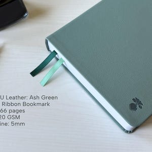 A green PU-leather notebook sits on a white wooden desk, showing 2 different color ribbon bookmarks that are of similar shade to the cover: teal and dark green. There is a fountain pen and ink bottle placed next to the notebook. Additional text in the image states that the notebook is an Ash Green color, has 366 pages, 120gsm paper, and is 5mm line.