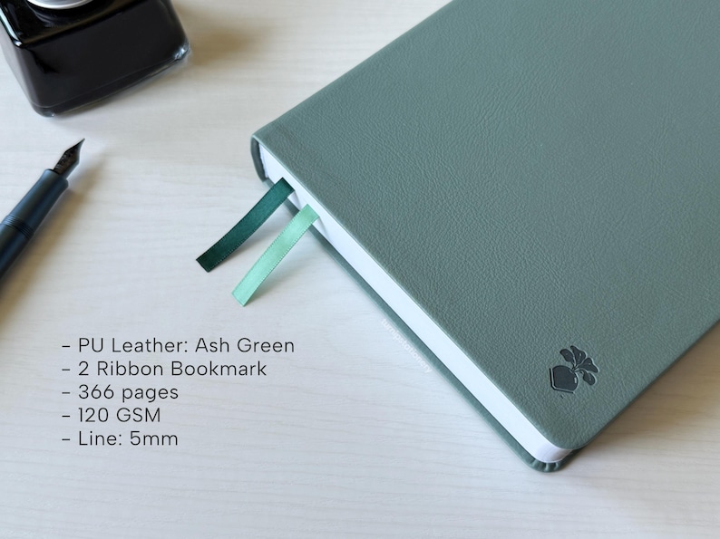 A green PU-leather notebook sits on a white wooden desk, showing 2 different color ribbon bookmarks that are of similar shade to the cover: teal and dark green. There is a fountain pen and ink bottle placed next to the notebook. Additional text in the image states that the notebook is an Ash Green color, has 366 pages, 120gsm paper, and is 5mm line.