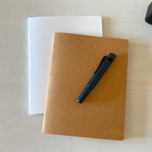 May include: A tan notebook with the number 404 embossed on the cover, layered over a white notebook. A dark blue fountain pen rests diagonally on the tan notebook, and a black ink bottle with a silver cap is positioned in the corner.