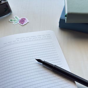 May include: A white notebook with a black pen lying on top of it. The notebook has a grid pattern and the pen is a fountain pen.